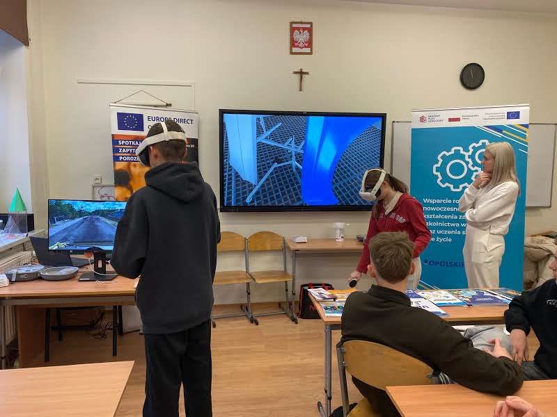 Widok na salę lekcyjną, w której uczniowie aktywnie korzystają z gogli wirtualnej rzeczywistości (VR) podczas zajęć. Na dwóch dużych monitorach widoczne są różne symulacje, w tym jazda samochodem i elementy konstrukcyjne. W tle znajduje się baner informujący o wsparciu na rzecz nowoczesnego kształcenia, a zajęcia prowadzone są w dynamicznej atmosferze.