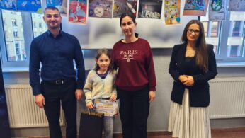 Zdjęcie pamiątkowe z wręczenia wyróżnienia. Od lewej stoją: mężczyzna w granatowej koszuli, dziewczynka trzymająca dyplom dla Julity Hohaisel i czarną torebkę prezentową, kobieta w bordowej bluzie z napisem „PARIS” oraz kobieta w okularach, czarnym żakiecie i białej plisowanej spódnicy. W tle wiszą prace plastyczne o tematyce kosmicznej.