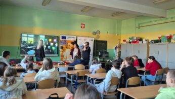 Szerokie ujęcie klasy lekcyjnej, w której uczniowie siedzą w ławkach i obserwują prezentację technologii VR. Na środku dwoje prowadzących nadzoruje osobę w goglach, a obraz z jej perspektywy jest wyświetlany na dużym ekranie interaktywnym. W tle widoczne są materiały edukacyjne „Europe Direct” oraz godło Polski.