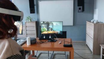 Zbliżenie na osobę z rudymi włosami, która ma na głowie białe gogle VR. Przed nią, na drewnianym biurku, znajduje się monitor wyświetlający symulację otwartej przestrzeni, głośniki oraz router. W tle widać białą tablicę suchościeralną i roślinę doniczkową.