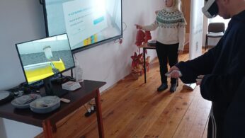 Zdjęcie przedstawia mężczyznę w goglach VR i z kontrolerami w dłoniach, który uczestniczy w wirtualnym szkoleniu. Obok stoi instruktorka wskazująca na duży ekran, na którym wyświetlane jest menu aplikacji oraz podgląd z gogli. Na mniejszym monitorze obok widać proces malowania natryskowego w środowisku cyfrowym.