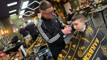 Mężczyzna w czarnej sportowej bluzie precyzyjnie strzyże nożyczkami włosy młodego klienta. Chłopiec siedzi w fotelu fryzjerskim, okryty czarną peleryną z żółtymi napisami „Barber Shop”. W tle widoczne jest stylowe, nowoczesne wnętrze salonu z wieloma stanowiskami pracy.