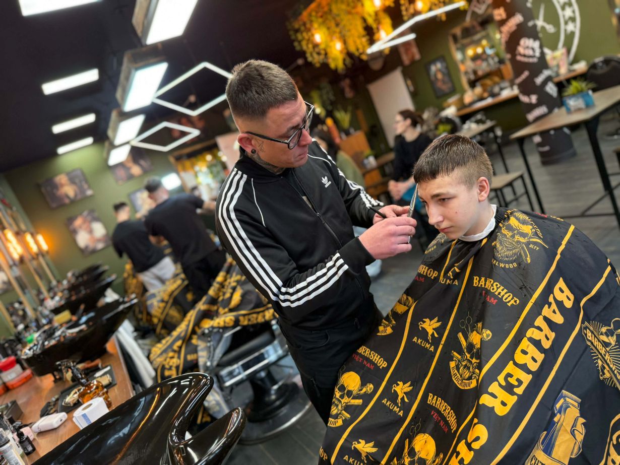 Mężczyzna w czarnej sportowej bluzie precyzyjnie strzyże nożyczkami włosy młodego klienta. Chłopiec siedzi w fotelu fryzjerskim, okryty czarną peleryną z żółtymi napisami „Barber Shop”. W tle widoczne jest stylowe, nowoczesne wnętrze salonu z wieloma stanowiskami pracy.