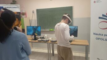 Szerokie ujęcie sali lekcyjnej z zieloną tablicą i godłem Polski. Dwóch uczniów korzysta z gogli VR przed monitorami ustawionymi na długim stole. Po lewej stronie widoczny jest baner 