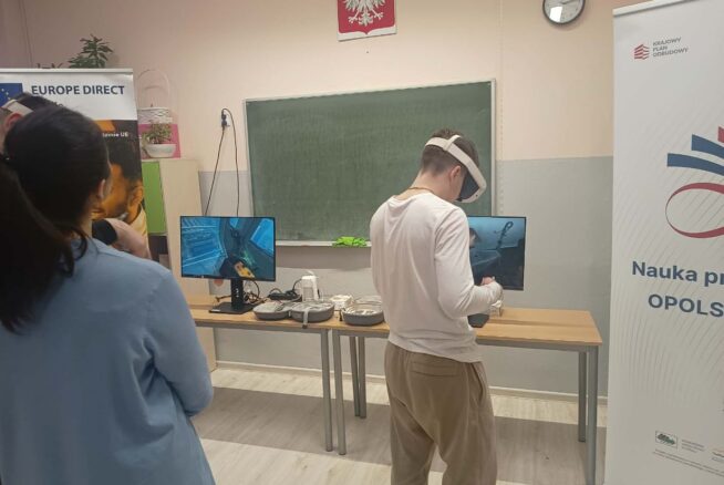 Szerokie ujęcie sali lekcyjnej z zieloną tablicą i godłem Polski. Dwóch uczniów korzysta z gogli VR przed monitorami ustawionymi na długim stole. Po lewej stronie widoczny jest baner "Europe Direct Opole", a po prawej roll-up z napisem "Nauka przez całe życie".