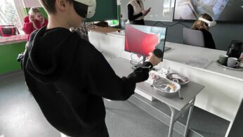 Zdjęcie przedstawia chłopca w czarnej bluzie z kapturem, który korzysta z gogli VR oraz kontrolera w jasnej sali lekcyjnej. Przed nim na biurku znajduje się monitor wyświetlający podgląd wirtualnego środowiska, a obok leży otwarte etui na sprzęt. W tle widoczna jest nauczycielka oraz duży ekran ścienny, na którym wyświetlany jest film z postacią kucharza. Pomieszczenie jest dobrze doświetlone światłem dziennym wpadającym przez okno.