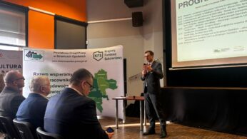 Zdjęcie przedstawia mężczyznę w ciemnym garniturze, który przemawia do zgromadzonych osób, trzymając mikrofon. W tle widoczny jest duży ekran z napisem 