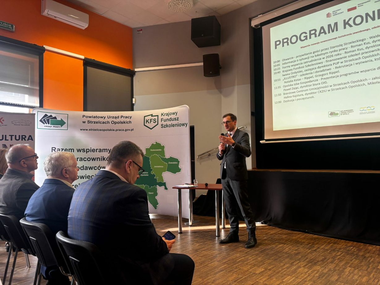 Zdjęcie przedstawia mężczyznę w ciemnym garniturze, który przemawia do zgromadzonych osób, trzymając mikrofon. W tle widoczny jest duży ekran z napisem "PROGRAM KONFERENCJI" oraz ścianka promocyjna Powiatowego Urzędu Pracy w Strzelcach Opolskich. Na pierwszym planie siedzą uczestnicy wydarzenia, z których jeden trzyma telefon. Sala konferencyjna ma nowoczesny wygląd z charakterystyczną pomarańczową ścianą.