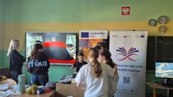 Grupa uczniów zgromadzona przed ekranem interaktywnym, na którym wyświetlany jest przód czerwonego samochodu w wirtualnym garażu. Po prawej stronie widać drugi, mniejszy monitor z innym widokiem oraz zegary ścienne. Na pierwszym planie widać stoły z naczyniami, co sugeruje przerwę lub poczęstunek podczas warsztatów.