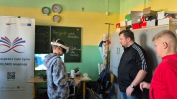 Uczeń w szarej bluzie z kapturem korzysta z gogli VR, podczas gdy instruktor w czarnej bluzie sportowej i uczeń w czerwonej bluzie przyglądają się jego działaniom. W tle widoczna jest tablica szkolna z napisami w języku niemieckim oraz baner programu edukacyjnego „Nauka przez całe życie”.