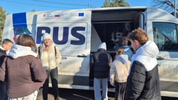 Uczniowie stoją na zewnątrz przy białym furgonie z napisem „EDUBUS”, rozmawiając z uśmiechniętą instruktorką w jasnej kurtce i czapce. Przesuwne drzwi pojazdu są otwarte, odsłaniając wnętrze wyposażone w grafikę z robotem i sprzęt technologiczny.
