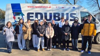 Grupa młodzieży wraz z opiekunką w futrzanej kamizelce pozuje do wspólnego zdjęcia na tle białego furgonu z dużym napisem „EDUBUS” i adresem strony internetowej www.opolskieLLL.pl. Akcja dzieje się na zewnątrz w słoneczny dzień, a na pojeździe widoczne są logotypy unijne oraz krajowe.