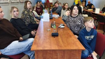 Widok na salę konferencyjną Powiatu Opolskiego, gdzie uczestnicy siedzą przy długich drewnianych stołach. Na ścianie widnieją tablice z portretami radnych różnych kadencji oraz godło Polski. W tle widać flagi Polski oraz żółto-niebieską flagę regionalną.