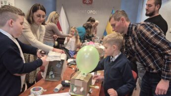 Ujęcie grupowe podczas pracy twórczej przy stole. Uczestnicy w różnym wieku współpracują przy budowie modeli z surowców wtórnych, takich jak kartony, butelki plastikowe i balony. W tle widoczny napis na ścianie 