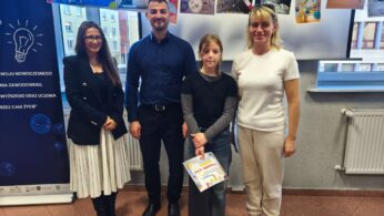 Uczestniczka konkursu, Mariia Ponomarenko, stoi z dyplomem w towarzystwie trojga dorosłych: kobiety w czarnym żakiecie, mężczyzny w niebieskiej koszuli oraz kobiety w białym t-shircie i jasnych spodniach dresowych. Grupa pozuje na tle wystawy rysunków i okna.