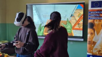 Zbliżenie na uczennicę w goglach VR, która za pomocą kontrolerów wchodzi w interakcję z wirtualnym środowiskiem. Na ekranie za nią widać podgląd aplikacji przedstawiający wnętrze magazynu z zielonymi rękawicami roboczymi. Obok stoi instruktorka obserwująca postępy dziewczyny.