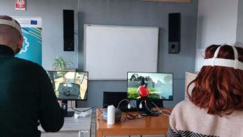 Dwie osoby siedzące przy biurku tyłem do obiektywu, korzystające z zestawów do rzeczywistości wirtualnej (VR). Na dwóch monitorach widoczne są różne symulacje: jedna przedstawia wnętrze kabiny pojazdu, a druga postać na zewnątrz. Na ścianie wiszą czarne głośniki oraz biała tablica.