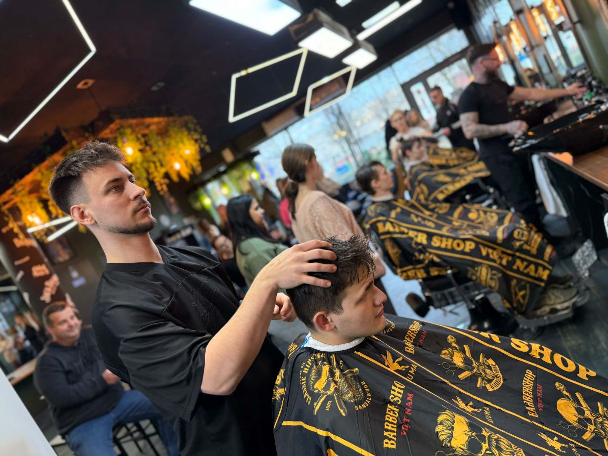 Fryzjer w czarnej koszulce modeluje dłońmi włosy młodego mężczyzny siedzącego w fotelu. Klient jest zabezpieczony peleryną fryzjerską z logo salonu, a w tle widać innych pracowników oraz osoby oczekujące. Wnętrze rozświetlają nowoczesne, geometryczne lampy sufitowe.