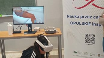Zbliżenie na uczennicę w goglach VR, która kuca przy stole, trzymając kontroler. Na monitorze przed nią wyświetlany jest symulowany obraz zabiegu medycznego lub badania nogi. Obok stoi roll-up z kodem QR i logotypami funduszy europejskich.