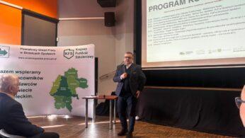 Fotografia ukazuje mężczyznę w ciemnej marynarce i okularach przemawiającego do publiczności w sali konferencyjnej. Za prelegentem znajduje się ekran z planem spotkania oraz baner z mapą powiatu strzeleckiego i logo Krajowego Funduszu Szkoleniowego. Z lewej strony widoczny jest profil siedzącego słuchacza. Pomieszczenie jest oświetlone naturalnym światłem wpadającym z boku.
