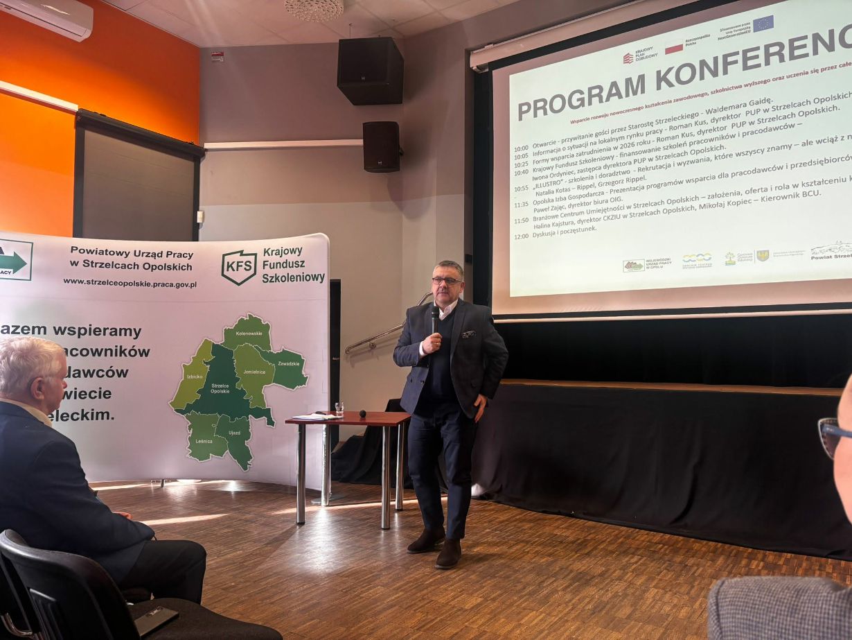 Fotografia ukazuje mężczyznę w ciemnej marynarce i okularach przemawiającego do publiczności w sali konferencyjnej. Za prelegentem znajduje się ekran z planem spotkania oraz baner z mapą powiatu strzeleckiego i logo Krajowego Funduszu Szkoleniowego. Z lewej strony widoczny jest profil siedzącego słuchacza. Pomieszczenie jest oświetlone naturalnym światłem wpadającym z boku.