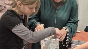 Zbliżenie na dłonie dwóch dziewczynek pracujących przy wspólnym projekcie. Jedna z nich okleja srebrną folią aluminiową konstrukcję, w której umieszczona jest plastikowa butelka. Na stole widać materiały plastyczne, w tym folię bąbelkową i kolorowe mazaki.