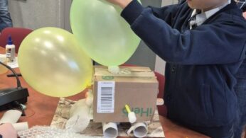 Chłopiec w granatowej bluzie stoi przy stole i za pomocą niebieskiego pisaka rysuje po jasnozielonym balonie. Balon jest przymocowany do kartonowego pudełka, które stanowi podstawę budowanego modelu. Na pierwszym planie widoczna jest butelka napoju oraz arkusz folii bąbelkowej.