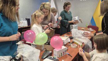 Szerokie ujęcie grupy dzieci i dorosłych pracujących przy długim stole. Uczestnicy budują modele z surowców wtórnych, takich jak kartony, balony, folia aluminiowa i bąbelkowa. Panuje atmosfera twórczego skupienia, a w tle widoczna jest flaga w barwach żółto-niebieskich.