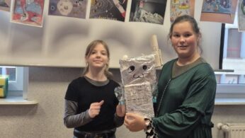 Dwie dziewczynki pozują z dużym modelem robota wykonanym z folii aluminiowej. Robot ma dorysowane oczy i uśmiech oraz ręce z plastikowych butelek. Tło stanowi tablica z kolorowymi rysunkami przedstawiającymi rakiety i planety.