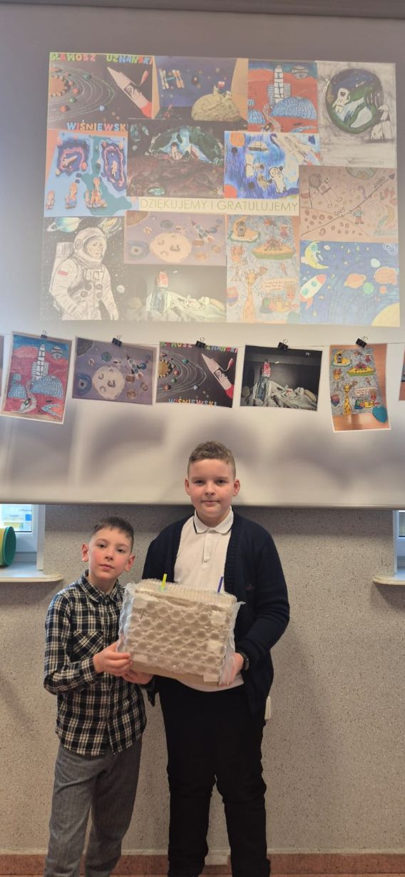 Dwóch chłopców stoi obok siebie, trzymając wspólnie duży model w kształcie sześcianu, który został w całości oklejony folią bąbelkową. W tle wiszą rysunki przedstawiające kosmos i astronautów.