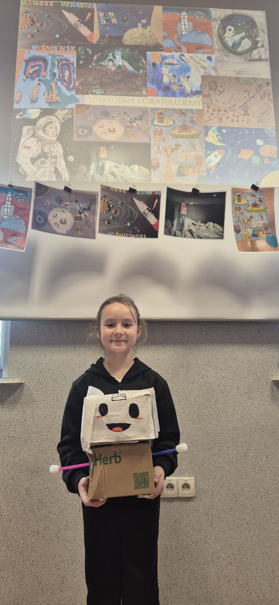 Dziewczynka w czarnej bluzie trzyma przed sobą model robota. Głowa robota wykonana jest z białego materiału z dorysowaną uśmiechniętą twarzą, a korpus stanowi brązowy karton z kolorowymi wypustkami po bokach.