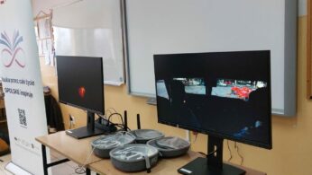 Stanowisko do nauki wirtualnej rzeczywistości z dwoma monitorami i akcesoriami ułożonymi na szkolnym biurku. Prawy monitor pokazuje widok z symulatora jazdy, prezentując plac manewrowy z czerwonymi elementami infrastruktury. Po lewej stronie kadru stoi baner z hasłem promocyjnym „Nauka przez całe życie – OPOLSKIE inspiruje”.