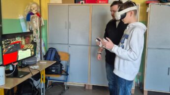 Instruktor w ciemnej bluzie pomaga uczniowi w białej masce VR obsługiwać kontrolery ręczne. Akcja dzieje się w rogu sali obok szafek, a na monitorze obok widać czerwony samochód w warsztacie. W tle widoczny jest model szkieletu człowieka z kolorowymi elementami.