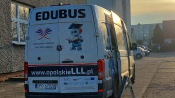 Widok z tyłu na biały samochód dostawczy marki Volkswagen Crafter, zaparkowany przed budynkiem. Na tylnych drzwiach widnieje napis „EDUBUS”, adres strony internetowej oraz grafika robota w birecie. W tle widać osiedle i zachodzące słońce.