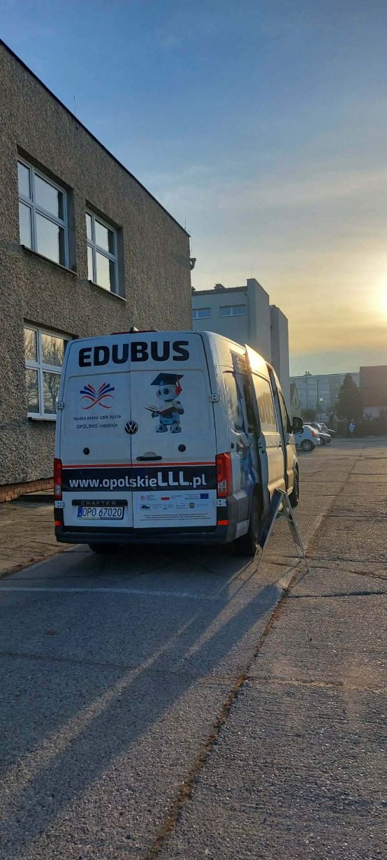 Widok z tyłu na biały samochód dostawczy marki Volkswagen Crafter, zaparkowany przed budynkiem. Na tylnych drzwiach widnieje napis „EDUBUS”, adres strony internetowej oraz grafika robota w birecie. W tle widać osiedle i zachodzące słońce.