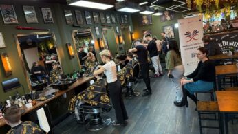 Szerokie ujęcie wnętrza barber shopu, w którym kilku fryzjerów jednocześnie obsługuje klientów przy długim blacie z lustrami. Na ścianach w oliwkowym kolorze wiszą liczne grafiki, a sufit zdobią drewniane i neonowe lampy. Po prawej stronie widać grupę osób obserwujących pracę oraz baner informacyjny.