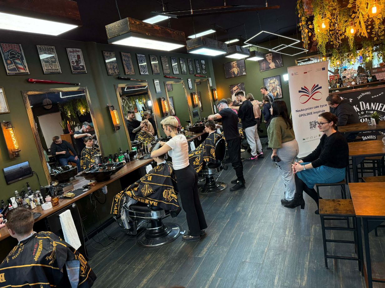 Szerokie ujęcie wnętrza barber shopu, w którym kilku fryzjerów jednocześnie obsługuje klientów przy długim blacie z lustrami. Na ścianach w oliwkowym kolorze wiszą liczne grafiki, a sufit zdobią drewniane i neonowe lampy. Po prawej stronie widać grupę osób obserwujących pracę oraz baner informacyjny.