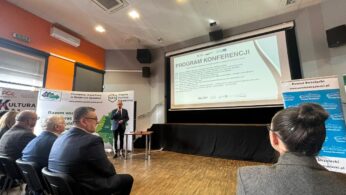 Szerokie ujęcie sali konferencyjnej z perspektywy uczestnika siedzącego w ostatnich rzędach. Na podwyższeniu stoi prelegent, a na dużym ekranie wyświetlany jest szczegółowy harmonogram konferencji. Po prawej stronie widoczna jest niebieska ścianka reklamowa Powiatu Strzeleckiego. Na suficie zamontowane jest profesjonalne oświetlenie sceniczne i klimatyzacja.