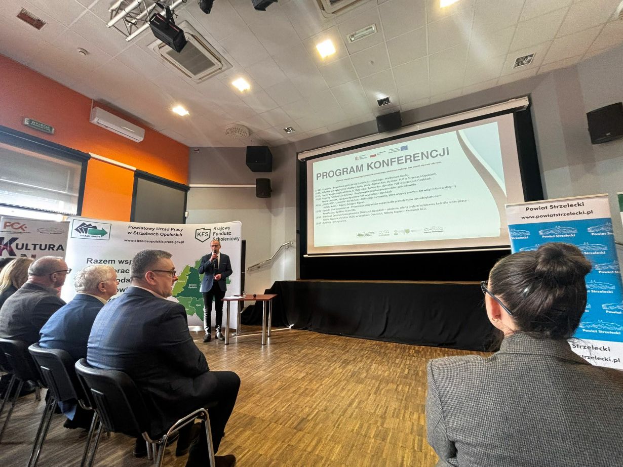 Szerokie ujęcie sali konferencyjnej z perspektywy uczestnika siedzącego w ostatnich rzędach. Na podwyższeniu stoi prelegent, a na dużym ekranie wyświetlany jest szczegółowy harmonogram konferencji. Po prawej stronie widoczna jest niebieska ścianka reklamowa Powiatu Strzeleckiego. Na suficie zamontowane jest profesjonalne oświetlenie sceniczne i klimatyzacja.