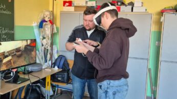 Zdjęcie przedstawia instruktora instruującego ucznia w brązowej bluzie z kapturem, jak poprawnie trzymać kontrolery VR. Na monitorze obok wyświetla się wirtualna scena przedstawiająca kuźnię lub warsztat rzemieślniczy. Obok monitora stoi szary pokrowiec na gogle oraz plecak.