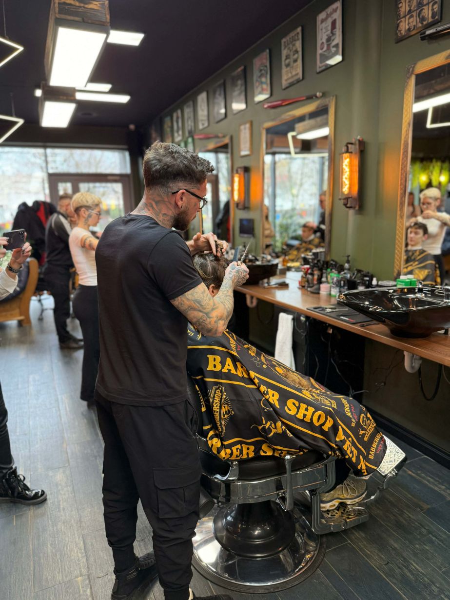 Fryzjer z licznymi tatuażami na ramionach strzyże włosy klienta przy użyciu nożyczek i grzebienia. Stanowisko pracy wyposażone jest w duże lustro w ozdobnej ramie, czarną umywalkę fryzjerską oraz profesjonalne kosmetyki. W tle widać fragmenty innych stanowisk i przeszklone wejście do lokalu.