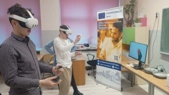 Dwóch uczniów stoi w sali lekcyjnej z założonymi goglami VR i kontrolerami w dłoniach. W tle widać różowe rolety w oknach oraz baner informacyjny 