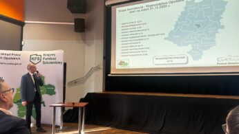 Mężczyzna w garniturze stoi na scenie i wskazuje na ekran z wyświetloną mapą oraz statystykami. Prezentacja dotyczy stopy bezrobocia w województwie opolskim na dzień 31.12.2025 roku. Obok prelegenta znajduje się biała ścianka z logo Urzędu Pracy. Zdjęcie zostało wykonane pod kątem, obejmując fragment widowni w dolnym rogu.