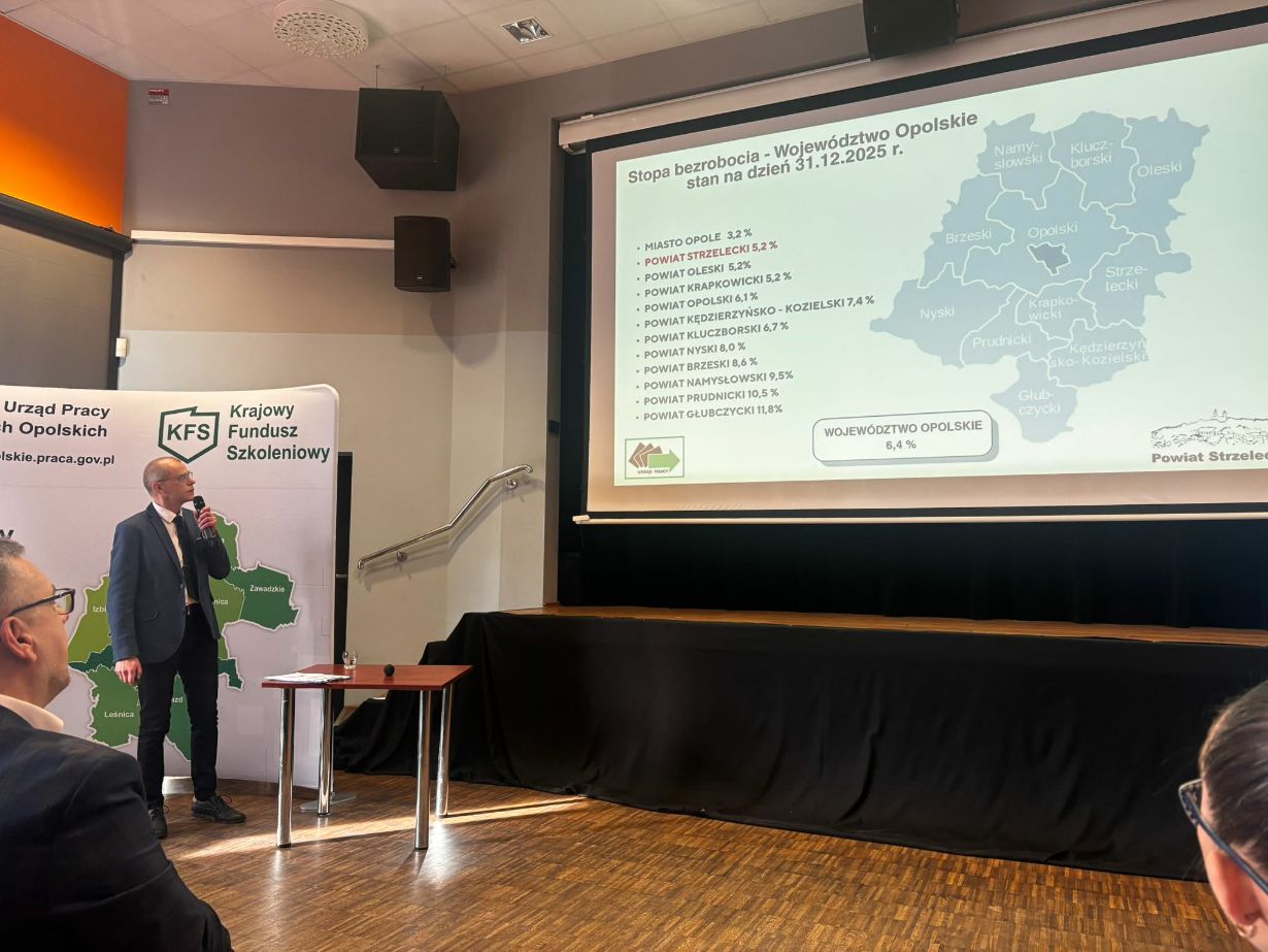 Mężczyzna w garniturze stoi na scenie i wskazuje na ekran z wyświetloną mapą oraz statystykami. Prezentacja dotyczy stopy bezrobocia w województwie opolskim na dzień 31.12.2025 roku. Obok prelegenta znajduje się biała ścianka z logo Urzędu Pracy. Zdjęcie zostało wykonane pod kątem, obejmując fragment widowni w dolnym rogu.