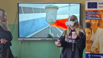 Uczennica w ciemnej bluzie z napisem korzysta z gogli VR, trzymając oba kontrolery przed sobą. Na dużym ekranie ściennym wyświetlany jest widok z aplikacji przedstawiający duży zbiornik przemysłowy w sterylnym pomieszczeniu. Z boku przygląda się jej blondynka z dłonią przy twarzy.