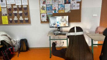 Perspektywa zza pleców uczennicy w goglach VR, która używa kontrolera. Na monitorze przed nią widać symulację medyczną/chemiczną. Na ścianie wiszą kolorowe rysunki dzieci z napisami „Chłopcy z Placu Broni”.