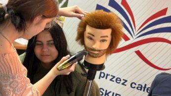 Zbliżenie na młodą kobietę, która ćwiczy strzyżenie brody maszynką na profesjonalnej głowie treningowej z rudymi włosami. Obok niej stoi inna dziewczyna, która z zaciekawieniem obserwuje proces nauki. W tle widoczny jest fragment białego baneru z kolorowym logotypem.