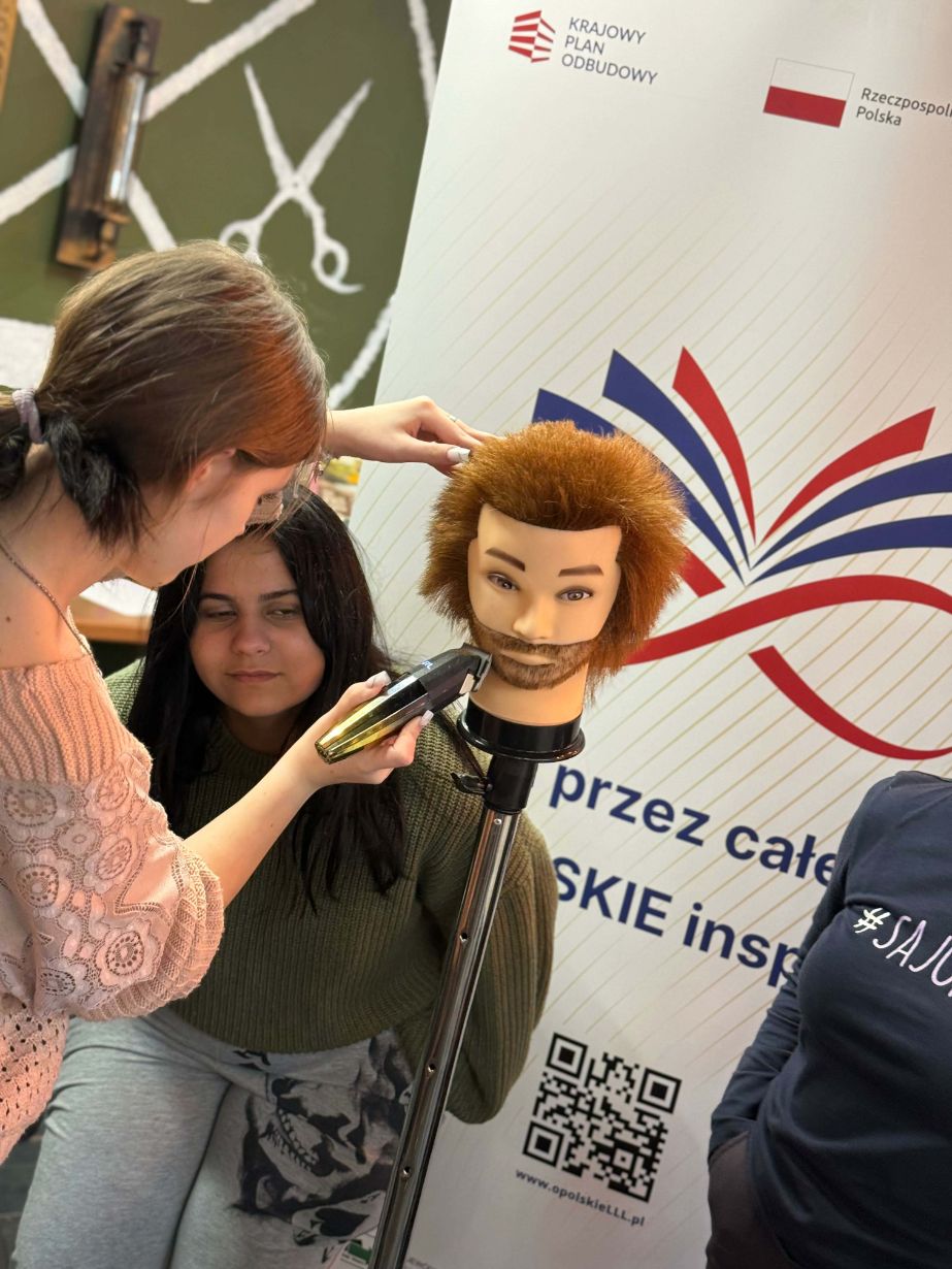 Zbliżenie na młodą kobietę, która ćwiczy strzyżenie brody maszynką na profesjonalnej głowie treningowej z rudymi włosami. Obok niej stoi inna dziewczyna, która z zaciekawieniem obserwuje proces nauki. W tle widoczny jest fragment białego baneru z kolorowym logotypem.