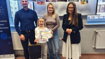 Uśmiechnięta dziewczynka, Julia Makuch, trzyma dyplom i czarną torebkę. Po jej bokach stoją: mężczyzna w niebieskiej koszuli, kobieta w beżowym golfie i jeansach oraz kobieta w czarnym żakiecie i białej spódnicy. Nad nimi wiszą rysunki przedstawiające planety i rakiety.