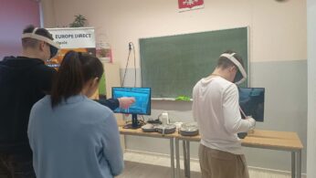 Ujęcie od tyłu na grupę osób zgromadzoną przy stanowiskach VR. Dwóch chłopców ma założone gogle i operuje kontrolerami, patrząc w stronę monitorów. Nad zieloną tablicą wisi zegar ścienny oraz godło Polski.